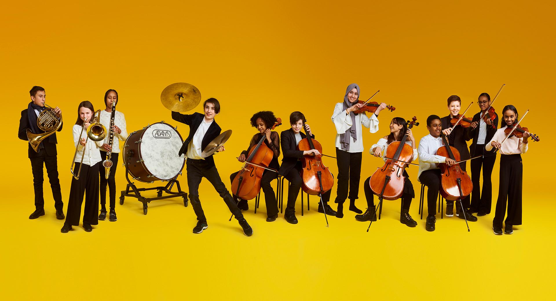 Barn i Kulturskolans orkesterskola/El sistema spelar på sina instrument mot en gul bakgrund.