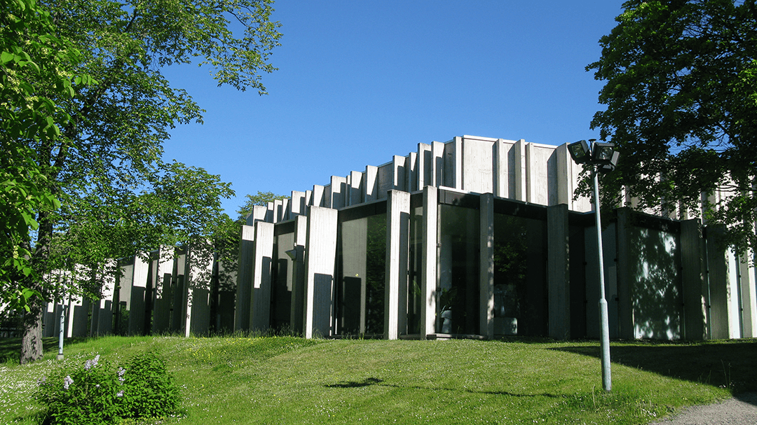 The brutalist facade of Berwaldhallen on a summer day with surrounding greenery and blue sky.