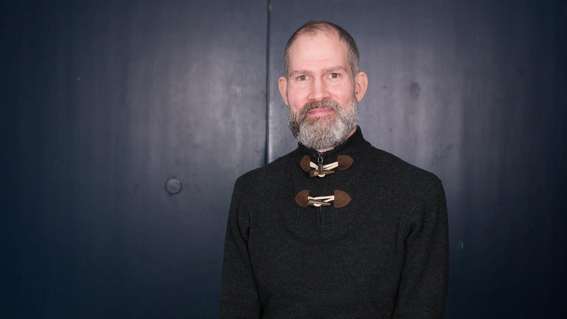 Joakim Schuster, Radiokören. Bild på man i grått skägg som tittar rakt in i kameran. Han ler och är klädd i en svart tröja med en arbetad knäppning.