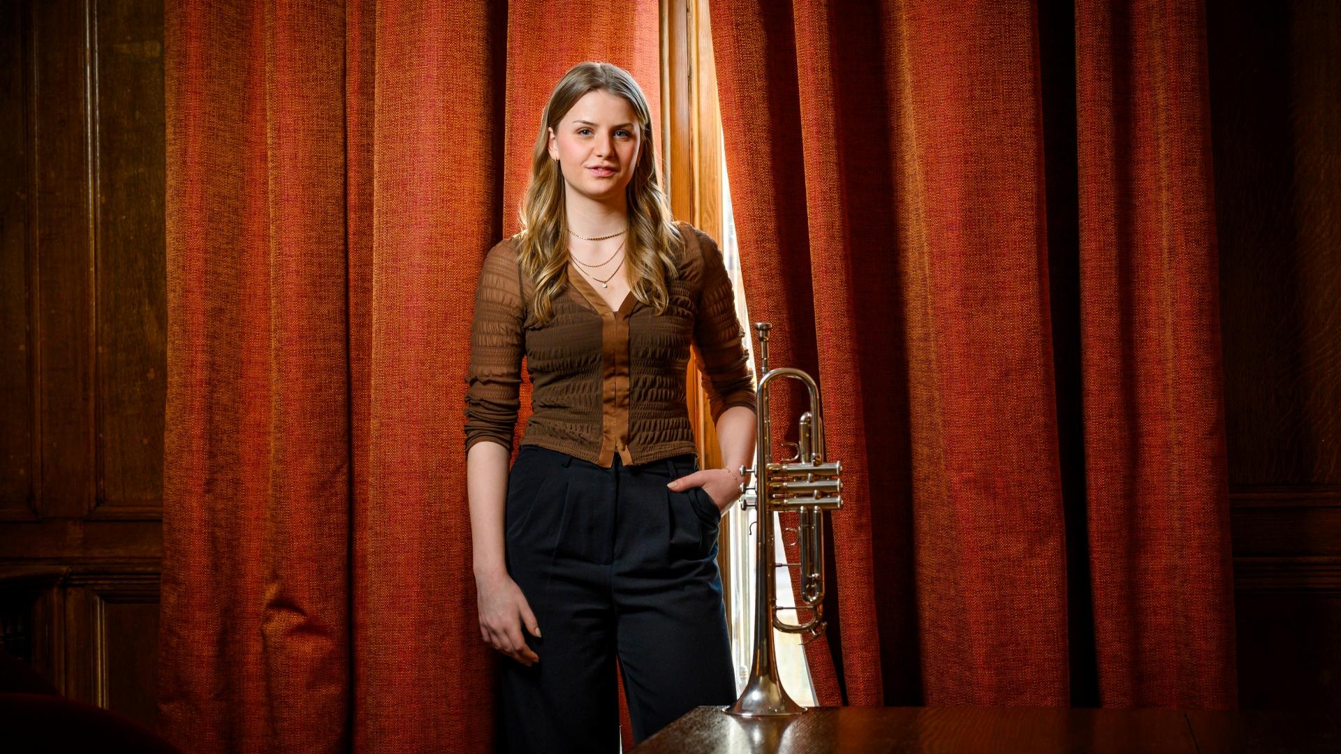 Ellinor Bengtson står framför ett rött sammetsdraperi i en brun blus och svarta byxor. Genom en glipa i draperiet ser tar sig ljus in. Hon har ställt sin trumpet på ett bord framför henne.