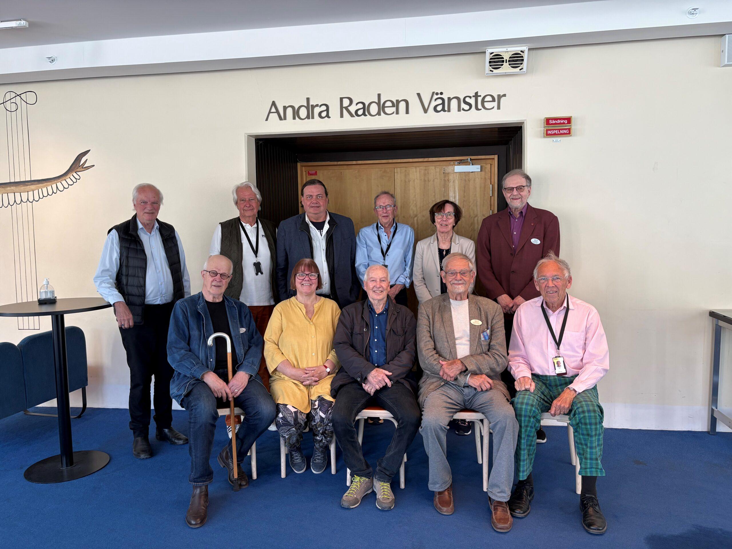 Vänföreningens styrelse står och sitter i två rader framför ingången med texten Andra Raden Vänster ovanför. De befinner sig i Berwaldhallen.