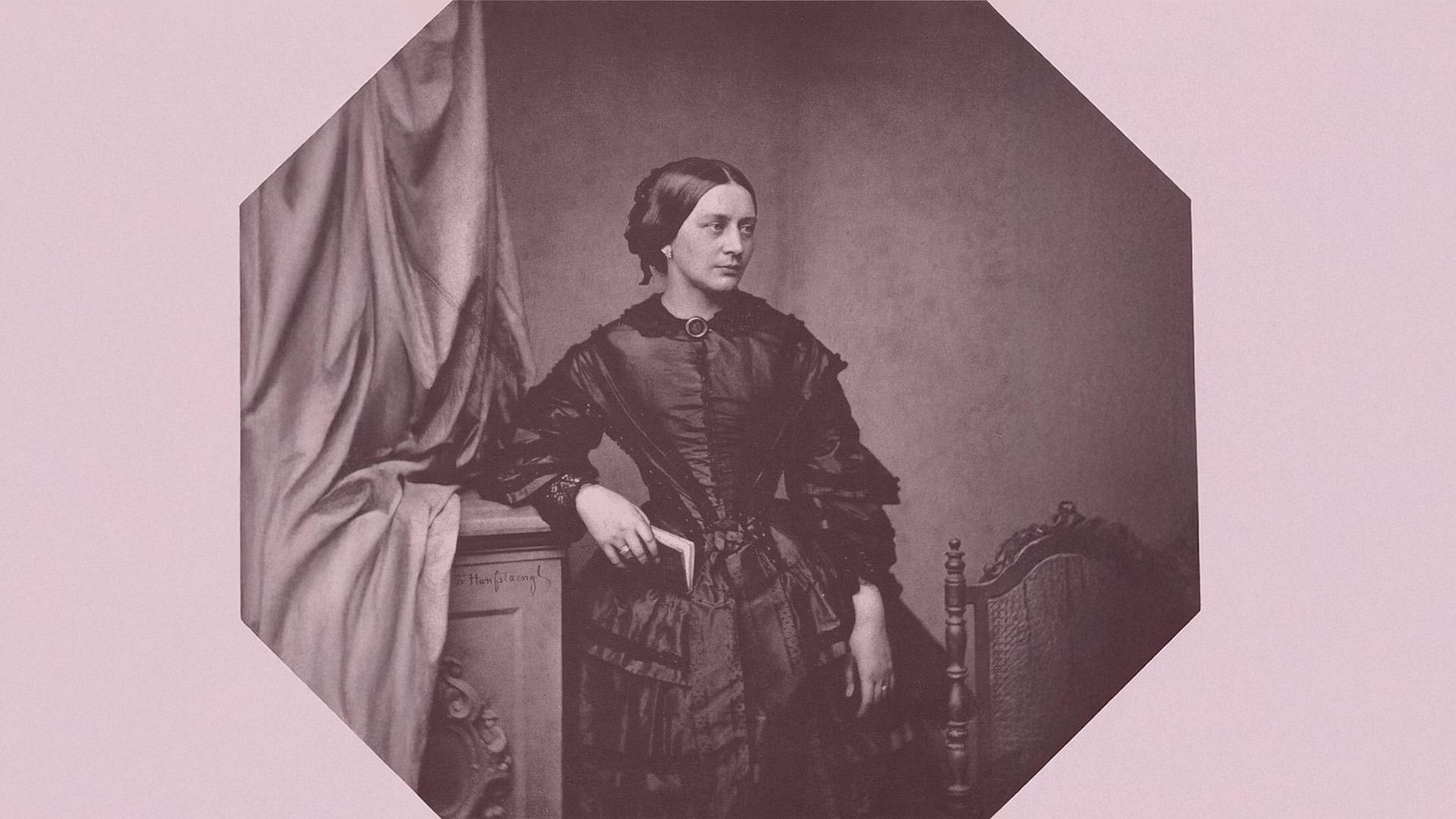 Stående Clara Schumann bär mörk klänning och lutar höger arm mot en byrå. Allvarsamt blickar hon åt sidan.